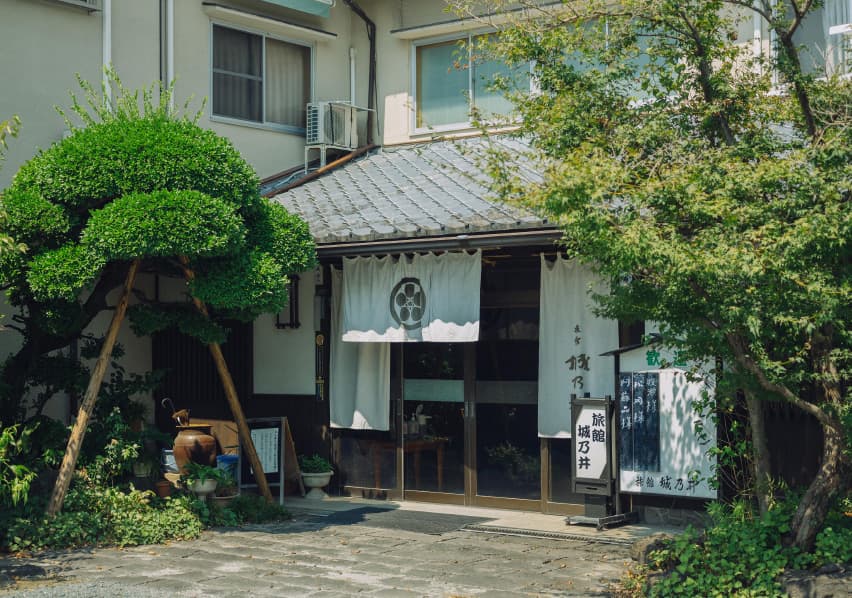 写真：綺麗に整えられた樹木の間に白いのれんがかかった玄関がある