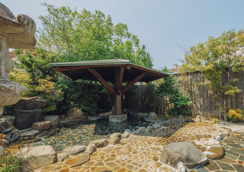 写真：板で周りが囲まれていて大きな木や日除けがある露天風呂